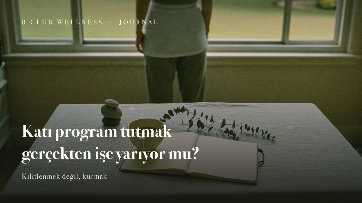 Keten örtülü masada üst üste dizili üç nehir taşı, bir fincan çay, açık boş bir defter ve kuru okaliptüs dalı; arkada pencerede sırtı dönük, uzakta duran bir kadın.